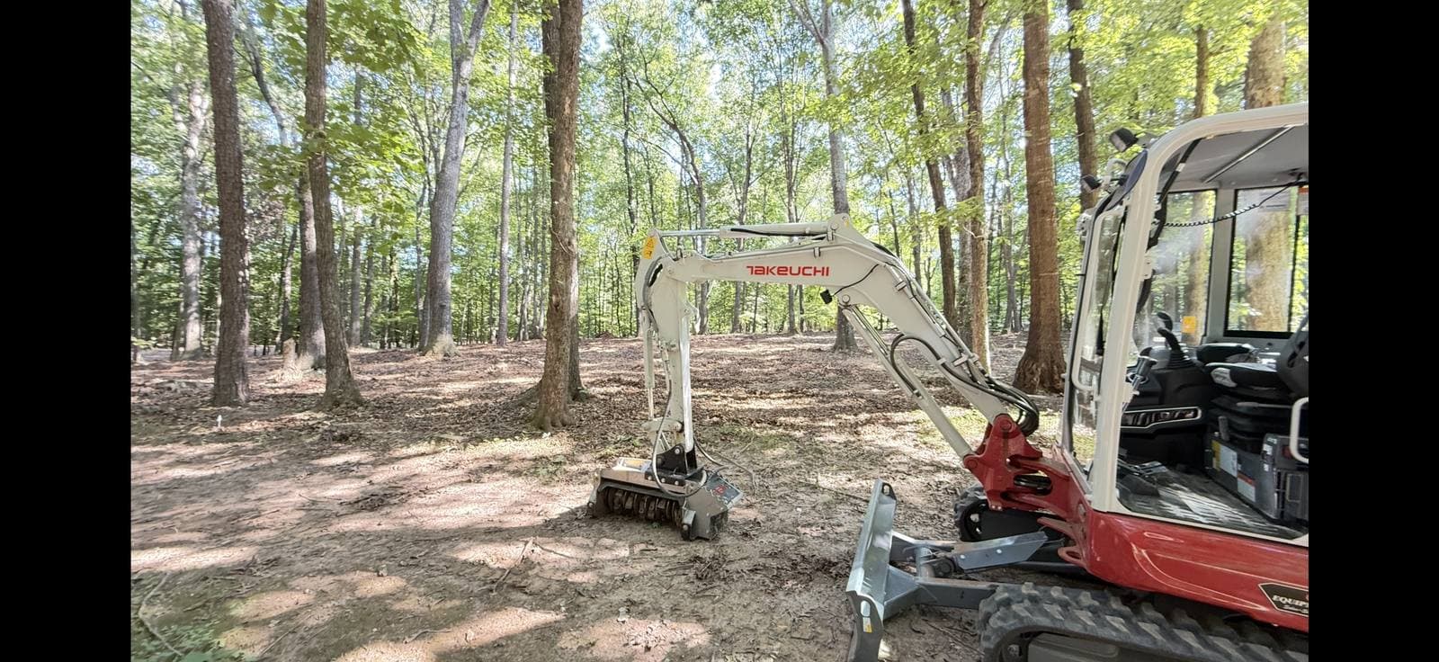 Mini excavator with a forestry mulcher clearing understory between hardwood trees on a wooded lot