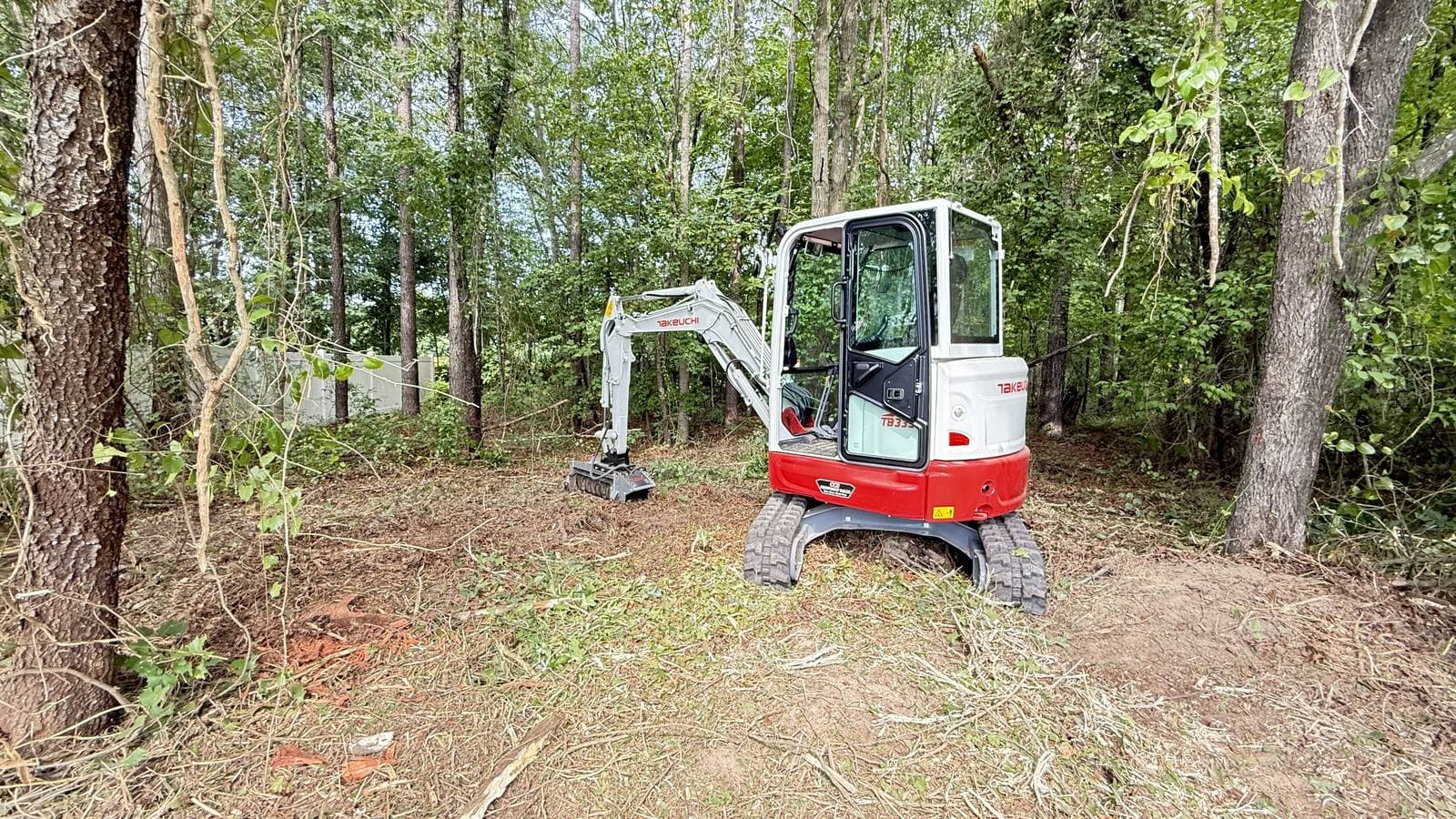 Takeuchi mini excavator thinning understory in a stand of hardwoods near a backyard fence