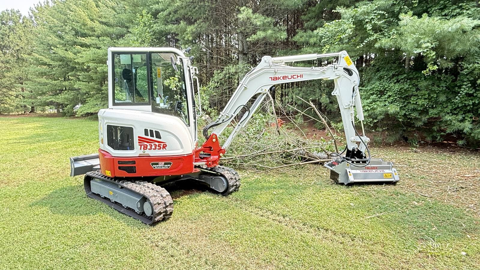 Takeuchi TB335R mini excavator with a forestry mulcher attachment parked on a lawn beside a pile of cut brush